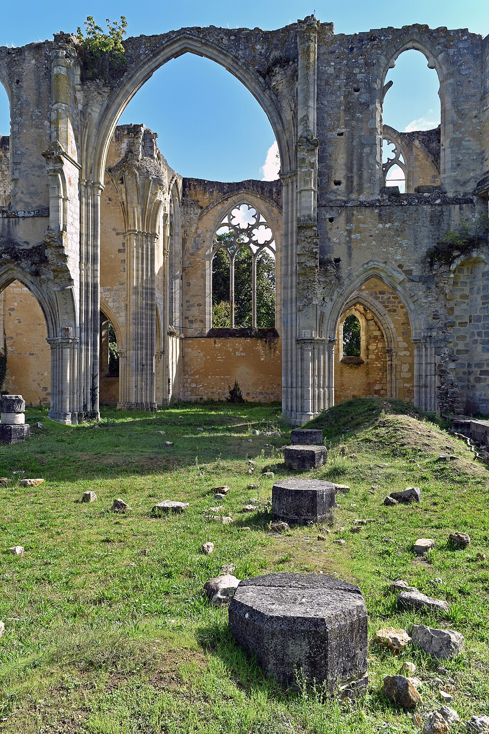 Murs de pierre conservant de grandes ouvertures et des arcs, sans toit visible au-dessus ; le ciel et la végétation apparaissent derrière. Au premier plan, des pierres et une base ronde sont entourées d’herbe.